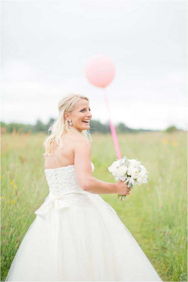 A bridal portrait taken outdoors in an open field with tall wild grass. The bride, a blonde woman with her hair loosely styled down, is turned slightly away from the camera and laughing over her shoulder. She wears a strapless ivory ballgown with a lace bodice, full tulle skirt, and a satin bow at the waist. She holds a compact bouquet of white peonies with dusty miller foliage, along with a large pink oversized balloon on a pink ribbon. The styling has a light, whimsical feel with a soft pastel palette. Medium portrait shot with a shallow depth of field, softly blurring the background.