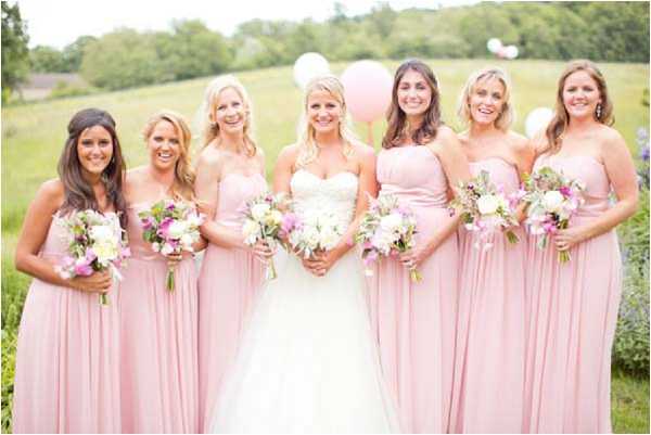 A bridal party portrait taken outdoors in a garden or lawn setting, featuring the bride and five bridesmaids posed in a line. The bride wears a strapless white ballgown with a beaded bodice and holds a white and cream bouquet of peonies and greenery. The bridesmaids wear floor-length strapless blush pink chiffon gowns and each holds a mixed bouquet of white, cream, and purple-pink blooms with greenery. Large white and pale pink balloons are visible in the background. The overall color palette is soft blush and white with a light, airy feel. Wide group portrait shot.