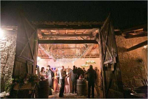 An evening reception scene photographed from outside looking into a rustic barn venue through large open wooden doors. Approximately 15-20 guests are gathered near the entrance, mingling and holding drinks, with several women wearing blush and mauve-toned dresses. The interior is decorated with draped fairy lights across the wooden beam ceiling and at least one chandelier visible in the background, creating warm ambient lighting. Wooden wine barrels are used as decorative elements and drink stations near the entrance, reinforcing a rustic, vineyard-style wedding aesthetic. The wide shot captures both the stone exterior walls and the warmly lit interior space.