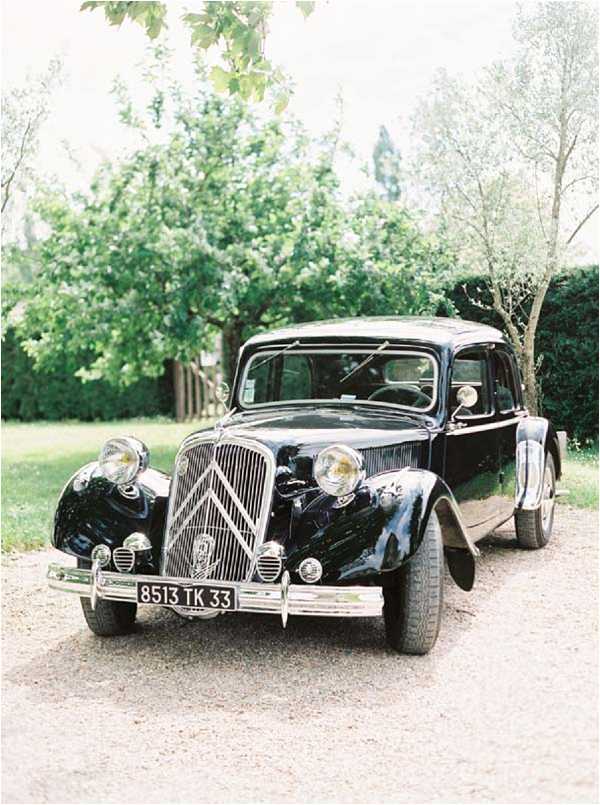 A vintage black Citroën Traction Avant, a classic French automobile, is parked on a gravel driveway as a wedding car. The vehicle features chrome headlights, a chrome front grille with the Citroën chevron emblem, and a French license plate reading 8513 TK 33, indicating the Gironde department. No people are visible in the frame. The shot is a straight-on wide portrait of the car, captured in a light, film-style aesthetic with soft exposure.