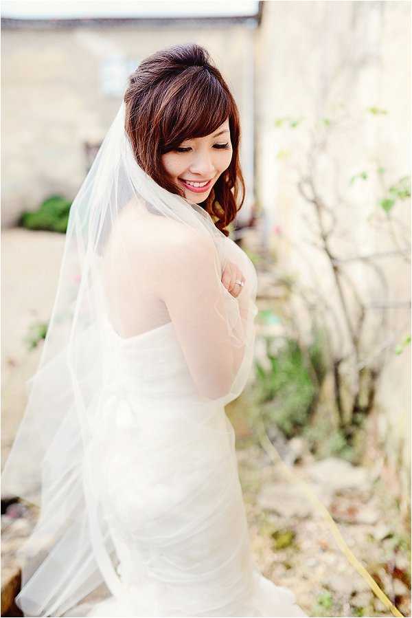 A bridal portrait of a single bride photographed outdoors against a softly blurred stone building backdrop. She wears a fitted ivory strapless gown with a draped or ruched bodice and a long plain-edge cathedral or elbow-length veil, and a solitaire ring is visible on her hand. Her dark auburn hair is styled in a loose half-up look with side-swept bangs, and she is glancing downward with a smile. The composition is a three-quarter-length portrait shot with a shallow depth of field, taken in natural daylight.