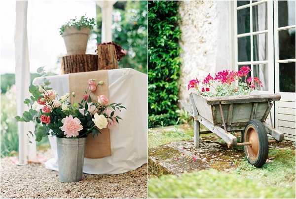 A two-image collage showing rustic outdoor wedding decor details at what appears to be a French country property. The left image is a close-up of a galvanized metal bucket filled with blush pink dahlias, cream lisianthus, small coral roses, and green foliage, placed beside a white linen-draped table with a burlap runner and wooden log stumps used as risers for terracotta pots. The right image shows a weathered wooden wheelbarrow filled with bright magenta and pink geraniums, positioned on a moss-covered stone surface beside the white-shuttered facade of a French country building. The overall styling theme is rustic-garden with a warm, natural palette of blush, coral, and magenta florals paired with raw wood and metal elements.