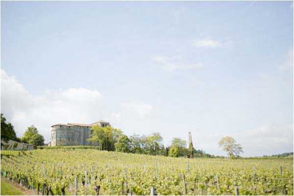 Wide landscape shot of a French vineyard estate, showing rows of vines in the foreground leading up to a stone chateau or winery building set on a gentle hill in the background. No people are visible in the image. The shot is taken from a low vantage point across the vineyard, emphasizing the scale of the property and its rural wine-country setting. Potential venue feature image.