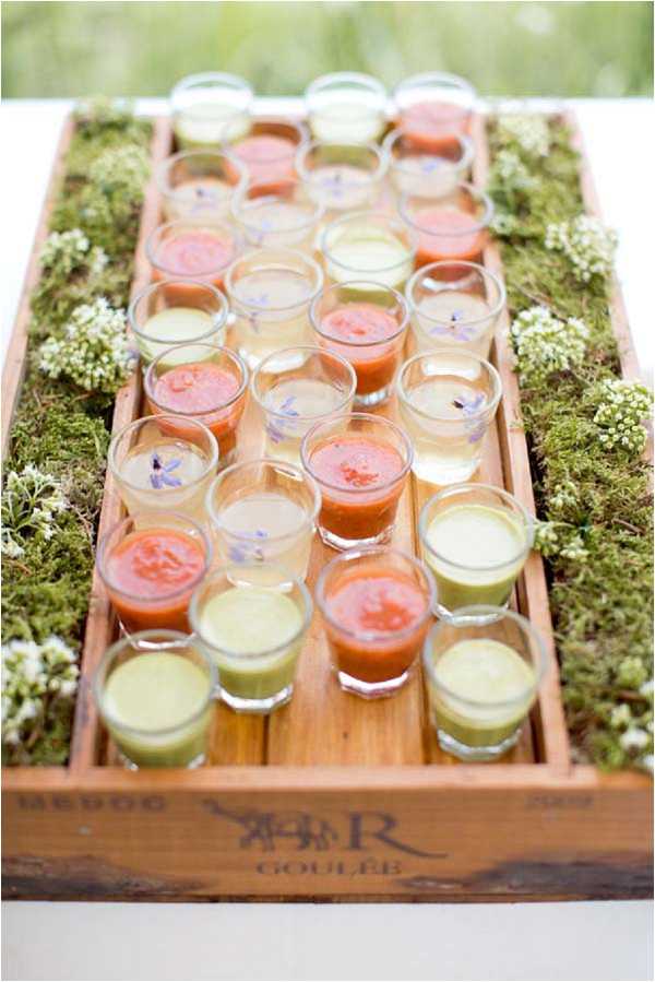 A close-up detail shot of a cocktail hour food presentation featuring rows of small shot glasses filled with three varieties of verrines — orange-red gazpacho, pale yellow or clear consommé garnished with blue borage flowers, and green vegetable-based soups — arranged in a rustic wooden wine crate stamped with 'GOULEE' branding. The crate is lined with green moss and small white wildflowers, creating a natural, garden-style presentation. The styling is rustic-organic, consistent with a French countryside wedding aesthetic.