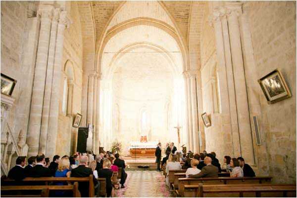 A wedding ceremony is taking place inside a Romanesque stone chapel with a barrel-vaulted nave and cream-colored limestone arches. Approximately 30–40 guests are seated in wooden pews on either side of a central aisle, facing the altar where the couple and officiant are standing. The altar area is backlit with warm natural light, and a small floral arrangement in warm tones is visible near the front. The wide-angle shot is taken from the rear of the nave, capturing the full length of the aisle and the architectural details including framed religious artwork on the side walls.