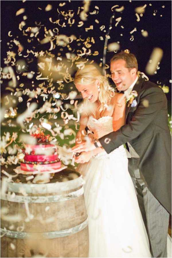 The bride and groom are cutting their wedding cake during an outdoor evening reception, with gold and cream paper confetti or ribbon streamers bursting around them mid-explosion. The bride wears a strapless ivory ballgown and the groom wears a charcoal grey suit with a satin lapel. The wedding cake is a small two-tiered red-hued cake decorated with berries and greenery, displayed on top of a rustic wooden wine barrel. Both the bride and groom are laughing and reacting with surprise to the confetti cannon. The setting appears to be an outdoor vineyard or winery venue, given the wine barrel display. The shot is a medium portrait taken at night with artificial lighting illuminating the couple and confetti against a dark background.