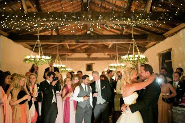 The bride and groom share their first dance at an indoor reception inside a rustic barn or agricultural outbuilding with exposed wooden beam ceilings. The ceiling is draped with warm white fairy lights, and two hanging chandeliers wrapped in greenery and white flowers serve as the central lighting fixtures. Approximately 30 guests surround the couple on the dance floor, many holding champagne glasses and wine glasses, with bridesmaids visible in coral/peach and pink dresses. The bride wears a strapless white ball gown and the groom is in a dark suit. The wide-angle shot captures the full room, the warm amber lighting from the fairy lights and chandeliers creating a cozy atmosphere in the rustic venue with stone or plaster walls.
