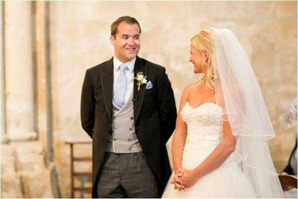 The bride and groom stand facing each other during what appears to be an indoor ceremony, both smiling. The setting features warm stone walls consistent with a French chapel or historic stone building. The groom wears a black morning coat with grey striped trousers, a silver waistcoat, pale blue tie, and a white and pale green boutonnière. The bride wears a strapless ivory ballgown with beaded or lace detailing on the bodice, paired with a two-tier elbow-length veil. The shot is a medium portrait framing both figures from approximately the knees up, with soft natural light illuminating the scene.