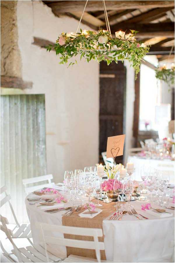 A reception table styling detail shot taken indoors at a rustic French venue with whitewashed walls, exposed wooden ceiling beams, and aged wooden doors. The round table is dressed in a white linen tablecloth with a burlap hessian runner, set with crystal wine glasses, silver cutlery, and place settings accented with small bright pink orchid blooms. Centerpieces include a low arrangement of hot pink hydrangeas in a mirrored vessel, white candles, and a small table number card held in a wire heart-shaped holder. Overhead, a circular hoop chandelier wrapped in trailing green ivy and small candles hangs from the ceiling, adding a botanical, rustic-chic styling element. The overall decor palette combines white, natural burlap, and hot pink florals in a relaxed rustic style.