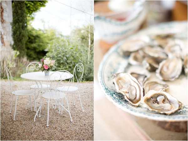 A split-image composition featuring two detail shots from what appears to be an outdoor wedding reception. On the left, a white wrought-iron bistro table and two matching chairs are arranged on a gravel courtyard, with a small floral centerpiece of pink and cream blooms in a silver vessel placed on the table. On the right, a close-up shot shows freshly shucked oysters served on a decorative blue-and-white ceramic plate, suggesting a cocktail hour or appetizer course. The overall styling is classic French with a romantic, light-toned palette of whites and soft pinks.
