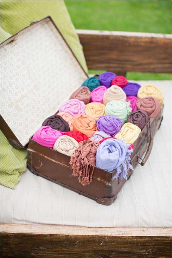 A close-up detail shot of a vintage brown leather suitcase, open and filled with tightly rolled pashmina or shawl wraps in a wide variety of colors including hot pink, magenta, teal, purple, orange, peach, mint, lavender, yellow, cream, brown, and dark plum. The suitcase is lined with a cream and gold patterned fabric and sits on a white cushioned wooden bench outdoors. A lime green folded blanket is visible to the left of the suitcase. This appears to be a guest comfort station setup offering wraps for an outdoor wedding reception or ceremony. The composition is a medium close-up shot at a slight angle.