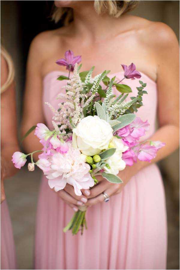 Close-up portrait of a bridesmaid holding a loosely arranged bouquet, with a second bridesmaid partially visible at the left edge of the frame. Both women wear strapless blush pink floor-length gowns. The bouquet features a white garden rose as the focal flower, accompanied by a blush peony, magenta sweet peas, deep purple clematis, white astilbe, green hypericum berries, lamb's ear, and eucalyptus foliage, creating a wild, garden-style arrangement. The styling is romantic and loosely structured with a soft pink and magenta color palette against the blush gowns.