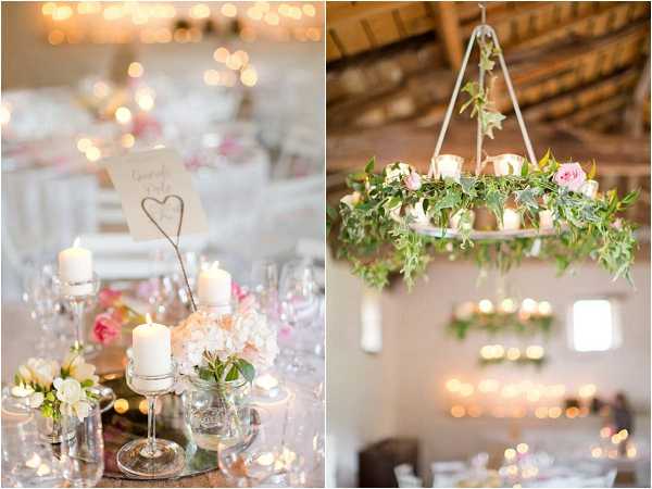 A two-panel detail shot of an indoor barn reception space decorated in a romantic rustic style with a soft pink and white palette. The left panel shows a close-up of a tablescape featuring white pillar candles on glass candleholders, small glass vases with blush hydrangeas and pink roses, tea lights, and a wire heart-shaped table number holder displaying a handwritten card. The right panel shows a white hoop chandelier suspended from a wooden-beamed ceiling, decorated with trailing green ivy, blush pink roses, and white flowers, with warm candlelight glowing in the softly blurred reception tables below. The overall decor palette combines blush pink, white, and greenery with warm ambient lighting throughout the barn space.