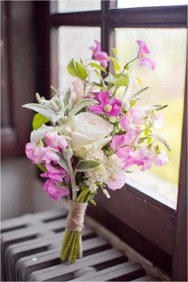 Close-up detail shot of a bridal bouquet resting on a dark metal radiator in front of a window with natural backlight. The loosely arranged, garden-style bouquet includes a cream ranunculus as the focal flower, pink and magenta sweet peas, magenta clematis blooms, small white hyacinth-like clusters, dusty miller foliage, and green leaves. The stems are wrapped with natural twine in a rustic style. The overall color palette is pink, magenta, cream, and soft grey-green, consistent with a romantic, wildflower-inspired aesthetic.