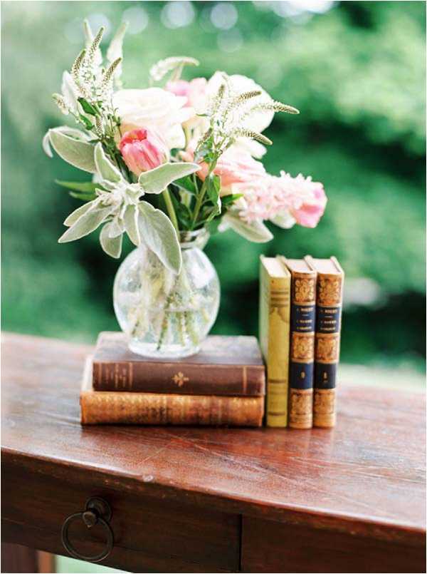 Close-up detail shot of a wedding decor vignette styled in a vintage literary theme. A small round clear glass bud vase holds a loose arrangement of blush pink ranunculus, coral tulips, dusty miller, and delicate white astilbe, placed atop two stacked antique leather-bound books with gold embossed spines in warm brown tones. Three additional aged hardcover books with tan and gold spines stand upright beside them, all arranged on a dark walnut wooden side table with a visible ring drawer pull. The overall palette is blush, coral, and soft green with warm brown vintage accents, consistent with a romantic, book-lover or garden-inspired wedding styling.