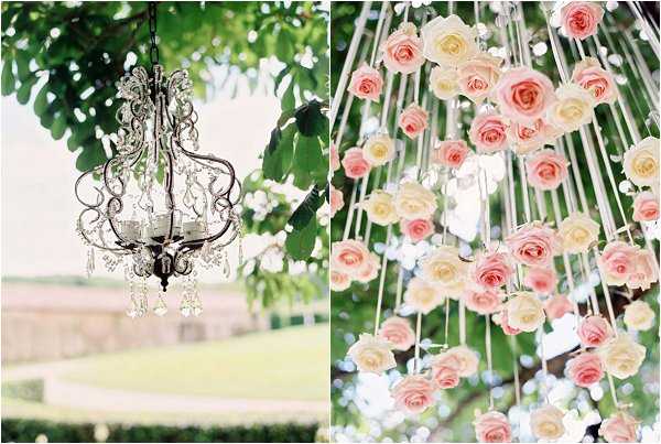 A split detail image showcasing two decorative elements from an outdoor wedding. On the left, a small ornate crystal chandelier with scrolled metalwork hangs from a tree branch, with a manicured garden visible in the soft background. On the right, a close-up of a suspended floral installation features blush pink and cream roses hanging individually on thin white ribbons or strings at varying heights, creating a cascading curtain effect against a backdrop of green foliage. The overall decor palette is romantic and classic, combining crystal accents with soft pink and ivory blooms. Both shots are close-up detail compositions with a soft, airy focus style.