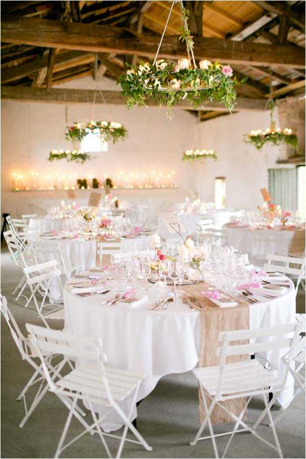 Indoor wedding reception setup inside a rustic barn venue with exposed wooden beam ceiling and whitewashed walls. Multiple round tables are dressed in white linen tablecloths with natural burlap or linen table runners, set with crystal glassware, silver cutlery, and small centerpieces featuring blush and hot pink roses with greenery in bud vases and pillar candles. White folding bistro-style chairs surround each table, and scattered pink flower petals decorate the place settings. Hanging floral chandeliers made from circular frames adorned with trailing greenery, cream and pink roses, and small candles are suspended from the ceiling beams, while a long ledge along the back wall is lined with rows of tea light candles creating a warm ambient glow. Wide shot capturing the full reception room with approximately six to eight tables visible. The overall decor palette is white, blush, and soft pink with natural greenery accents in a rustic-romantic style. Potential venue feature image.
