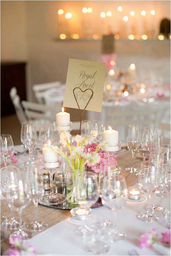 A close-up detail shot of a wedding reception table centerpiece in an indoor venue. The centerpiece features a glass vase of pink and white sweet peas and tulips, surrounded by white pillar candles on glass candleholders and scattered tea light candles. A gold card table name holder with a wire heart detail displays the table name 'Royal Ascot' in calligraphy script. The table is set with multiple crystal wine and champagne glasses, white linen, and a neutral-toned table runner. Loose pink sweet pea blooms are scattered along the table. The decor palette is soft pink, white, and gold with a classic, romantic styling. In the background, additional candlelit tables are visible along with a row of glowing candles or fairy lights creating a warm, amber-toned atmosphere in the reception space.