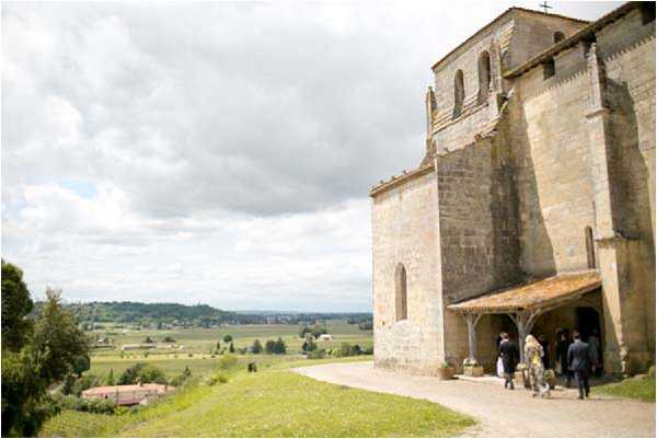 Guests arrive at a Romanesque stone chapel set on an elevated hilltop site, with a sweeping view of a rural French valley stretching into the distance below. The wide exterior shot captures approximately four to five guests in formal attire, including one figure in a yellow outfit, walking toward the arched portico entrance of the chapel. The architecture features thick limestone masonry, a tiled roof portico, and arched windows typical of medieval French religious buildings. Potential venue feature image.