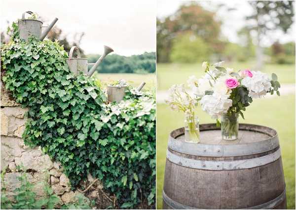 A split image showing two detail shots from an outdoor rustic-style wedding. On the left, galvanized metal watering cans are arranged on an ivy-covered stone wall, used as decorative or functional elements. On the right, a wooden wine barrel serves as a drinks station or decor stand, topped with two glass jar vases holding a loose arrangement of white hydrangeas, hot pink roses, and eucalyptus foliage. Both shots are close-up detail compositions taken in an outdoor garden or countryside setting, consistent with a rustic French countryside wedding aesthetic.