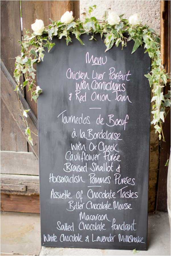 A close-up detail shot of a chalkboard wedding menu sign leaning against a rustic wooden door. The menu, written in white and pink chalk handwriting, lists a three-course dinner: Chicken Liver Parfait with Cornichons and Red Onion Jam, Tournedos de Boeuf à la Bordelaise with Ox Cheek, Cauliflower Purée, Braised Shallot and Horseradish Pommes Purées, followed by an Assiette of Chocolate Tastes including Bitter Chocolate Mousse, Macaroon, Salted Chocolate Fondant, and White Chocolate and Lavender Milkshake. The top of the chalkboard is decorated with a garland of trailing ivy and cream/white roses. The overall styling is rustic and relaxed, consistent with a French countryside wedding setting.