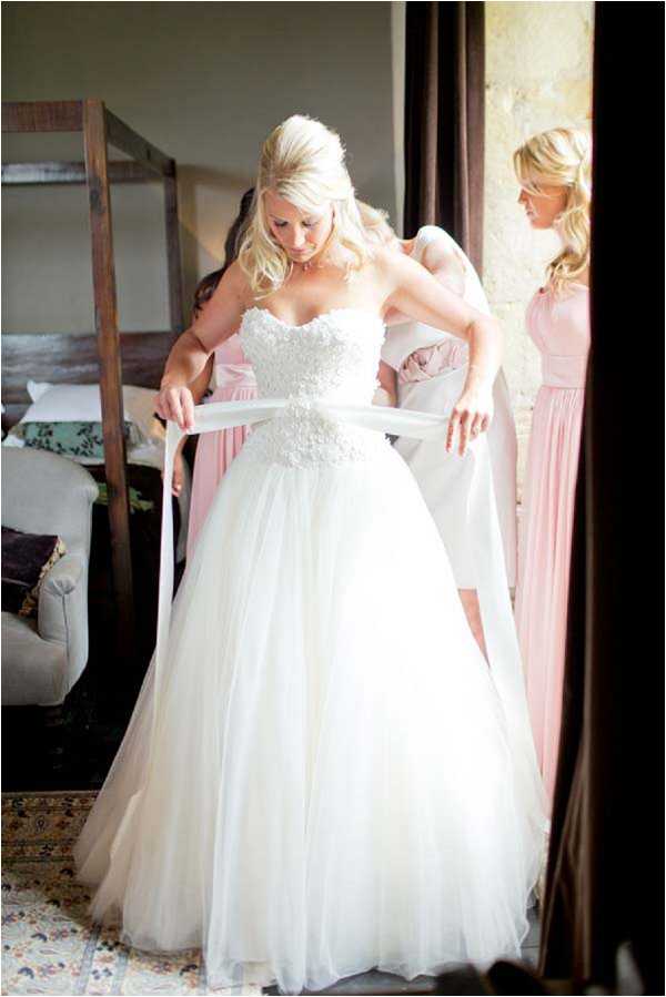 A getting-ready scene captured indoors, showing a bride being assisted by two bridesmaids as a white sash or ribbon is tied around her waist. The bride wears a strapless ball gown with a lace and floral appliqué bodice and a full tulle skirt, with her blonde hair styled in an updo. The two visible bridesmaids are dressed in floor-length blush pink gowns. The room features a rustic wooden mirror frame, a patterned area rug, and stone wall details visible in the background, suggesting a chateau or mas-style venue. The shot is a medium portrait taken from a doorway, with soft natural light illuminating the scene.