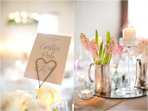 Close-up detail shots of wedding reception table decor presented as a diptych. On the left, a kraft paper table name card reading 'Cartier Polo' is held in a wire heart-shaped card holder, positioned among cream garden roses with soft bokeh lighting in the background. On the right, a silver/pewter pitcher holds pink hyacinths and white tulips as a centerpiece, accompanied by a tall glass pillar candle holder with a white candle, a small glass tealight holder, and additional silver bud vases, all arranged on a mirrored tray with a taupe table runner. The overall decor palette combines blush pink, cream, and silver metallic tones in a romantic classic style.