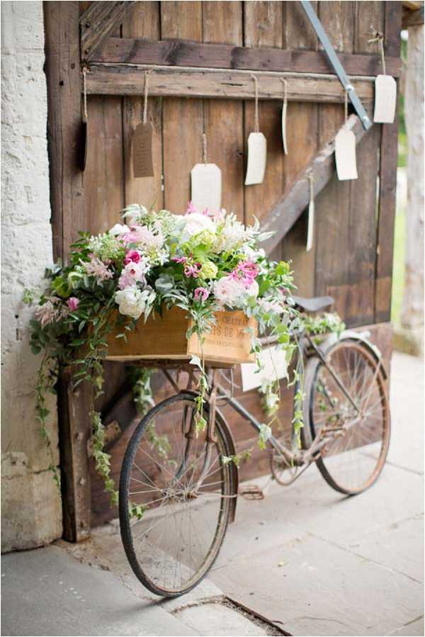 A close-up detail shot of a vintage rusty bicycle used as a wedding decor prop, leaning against large weathered wooden barn doors. The bicycle's front basket holds a wooden wine crate filled with an overflowing floral arrangement of pink peonies, hot pink ranunculus, cream roses, and trailing green ivy and foliage. Behind the bicycle, cream luggage-tag style escort cards are suspended from a horizontal rod by twine in front of the wooden doors. The overall styling is rustic bohemian, combining repurposed vintage items with lush garden-style florals.