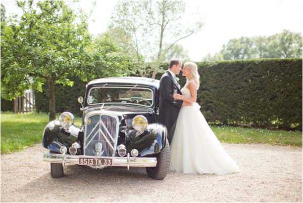 A couple poses together in front of a vintage black Citroën Traction Avant with a French license plate (8513 TK 33), parked on a gravel driveway. The bride wears a strapless white ballgown with a full tulle skirt and holds a champagne flute, while the groom is dressed in a dark suit; they stand close together facing each other. The setting is an outdoor garden area with a tall trimmed hedge and trees in the background. The shot is a medium-wide portrait composition with soft, bright natural lighting.