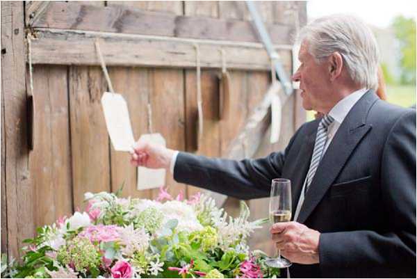 A male guest in a dark suit with a grey striped tie holds a champagne flute in one hand and examines a small hanging tag with the other, at what appears to be an outdoor seating chart or escort card display attached to a rustic wooden wall or barn door. The cards are suspended from a horizontal rod with string. In the foreground, a large floral arrangement features pink roses, white astilbe, green hydrangea, and eucalyptus foliage. The setting is rustic in style, and the shot is a medium portrait taken from a slight angle.