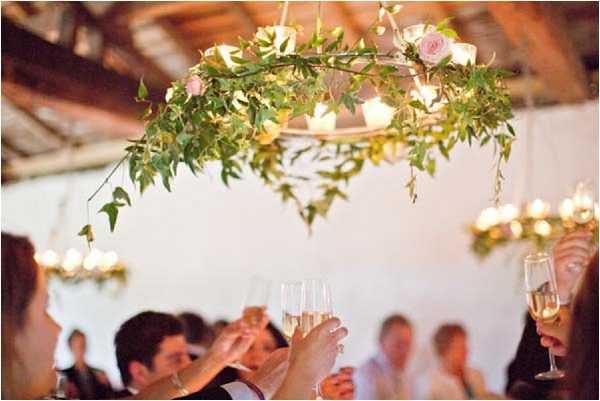Guests raise champagne flutes in a toast during what appears to be a wedding reception, with the foreground hands and glasses in sharp focus while the seated crowd is softly blurred in the background. The setting is an indoor rustic venue with exposed wooden ceiling beams and white walls. Hanging above the space is a circular floral chandelier decorated with trailing green foliage and small blush pink roses, accented with warm candlelight bulbs; additional candle chandeliers are visible in the background. The overall decor palette is soft and natural, combining greenery, blush tones, and warm ambient lighting in a style that reads as relaxed rustic-romantic.