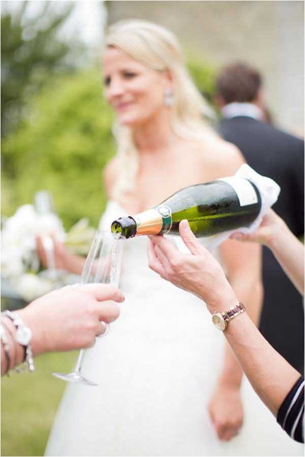 A close-up shot of champagne being poured from a green bottle with a gold foil cap into a tall champagne flute during an outdoor cocktail hour. In the foreground, one hand holds the flute while another pours, with a gold watch and bracelet visible on the wrist of the person pouring. In the soft-focus background, a blonde bride in a white strapless gown is visible smiling, holding what appears to be a white bouquet, with at least one other guest in a dark suit partially visible beside her. The setting appears to be an outdoor garden or grounds area.