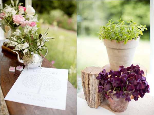 A split close-up detail shot showing two styled vignettes from what appears to be an outdoor or semi-outdoor wedding with a rustic, garden-inspired theme. On the left, a vintage silver teapot holds a loose arrangement of white astilbe, pink roses, and greenery, placed on a dark wood surface alongside a printed wedding program featuring a tree illustration and the names Stan and Nicky. On the right, two terracotta pots — one containing fresh green herbs and one filled with deep purple oxalis flowers — are displayed on a weathered wood log slice used as a natural riser. The overall decor palette combines silver, blush pink, deep purple, and natural wood tones, suggesting a romantic rustic styling approach.