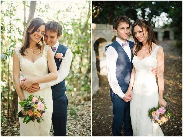 A diptych of two couple portraits taken outdoors at what appears to be a French property with stone archway ruins visible in the right image. The bride wears a fitted ivory lace gown with a sweetheart neckline and holds a loose bouquet of peach roses, purple flowers, and greenery. The groom wears a navy waistcoat over a white shirt with a lilac bow tie. In the left portrait, the couple share a close, intimate pose in a sun-dappled wooded area; in the right portrait, they stand facing the camera in front of the stone archway ruins. The styling has a relaxed, boho-romantic feel with a soft pastel color palette. Both images are medium portrait shots with warm, natural light.