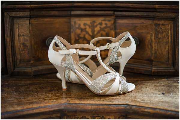 Close-up detail shot of a pair of bridal heels displayed on a dark carved wooden surface, likely an antique dresser or cabinet. The shoes are ivory satin t-strap heels with silver and gold glitter embellishment along the straps and toe band, featuring an ankle buckle closure and an open toe design. The pairing of ivory satin and metallic glitter detailing gives the shoes a classic bridal aesthetic. The carved wood background provides a rich, dark contrast to the light-colored footwear.