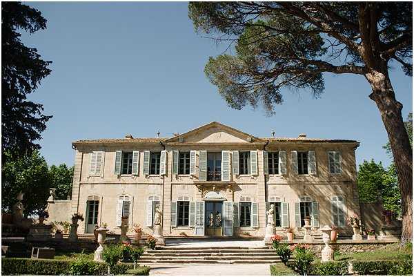 Wide exterior shot of a classic French chateau featuring a symmetrical two-story stone facade with pale blue-grey shutters, a triangular pediment, and a grand central staircase leading to the main entrance. The building is flanked by decorative stone urns, classical statuary, and potted flowering plants in terracotta tones along the steps and balustrades. No people are visible in the frame; the image serves as a venue overview shot taken in bright daylight. Potential venue feature image.