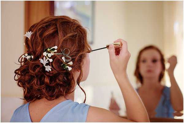 A getting-ready scene showing a woman with auburn hair styled in a loose updo adorned with small white floral hair accessories and a delicate wire bow detail, applying makeup with a brush while looking into a mirror. The shot is taken from behind, with her reflection visible in the mirror in the background. She is wearing a pale blue sleeveless dress or top, suggesting she may be a bridesmaid. The composition is a close-up portrait focusing on the hair styling detail, captured indoors in a softly lit room.