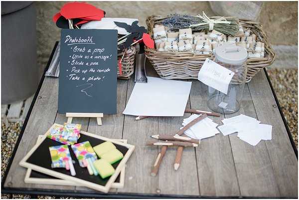 A close-up detail shot of an outdoor wedding activity table set on a weathered wooden surface. The table features a photobooth station with a small chalkboard sign on a miniature easel reading 'Photobooth – Grab a prop, Write a message, Strike a pose, Pick up the remote, Take a photo,' alongside red and black prop cutouts on sticks. Additional items include small framed chalkboards with colorful chalk pieces, rustic twig-style pencils, blank white cards, a glass jar, and a large wicker basket filled with individually wrapped lavender bundle favors tied with twine. The overall styling is rustic with natural and handmade elements.