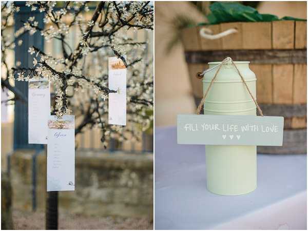 A side-by-side detail shot of two rustic-style wedding decor elements at an outdoor venue. On the left, white paper seating chart cards are tied with twine and hung from the branches of a flowering tree with small white blossoms, creating an informal escort card display. On the right, a mint-green painted metal milk churn sits on a pale blue linen-covered table, with a small wooden sign hanging from jute rope reading 'Fill your life with love' with three small heart motifs; a wooden barrel is partially visible in the background. The overall decor palette combines mint green, pale blue, and natural wood tones in a rustic, countryside styling.