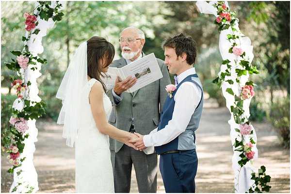 An outdoor wedding ceremony taking place beneath a white floral arch decorated with pink roses and trailing green ivy. The bride, wearing a sleeveless ivory lace dress and a cathedral-length veil, holds hands with the groom, who is dressed in navy trousers, a white shirt, and a grey waistcoat with a pink rose boutonnière. An elderly male officiant in a grey suit holds an open ceremony booklet and reads aloud. The setting is a tree-lined garden pathway. The image is a medium wide shot capturing all three figures with the arch framing the couple.