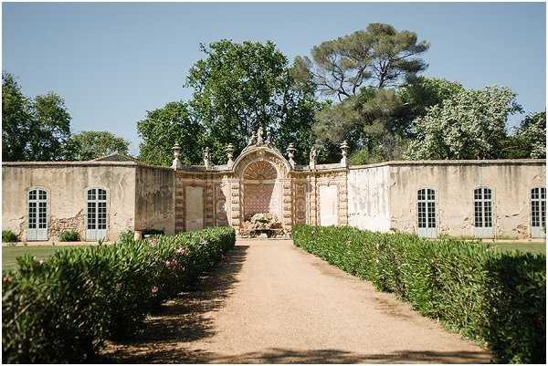 Wide shot of a formal French estate garden pathway leading to an ornate stone gateway with a decorative arched niche, sculpted figures along the roofline, and symmetrical wings featuring pale blue-grey shuttered arched windows. The gravel path is flanked by neatly trimmed low hedgerows. No people are visible in the frame. Potential venue feature image.