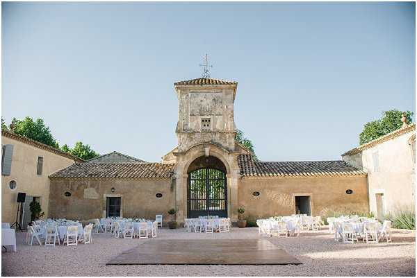 A wide-angle outdoor shot of a wedding reception setup in the courtyard of a historic Provençal mas or domaine, featuring ochre-rendered stone buildings with terracotta tile roofs and a central arched gateway topped with a bell tower and iron cross. Multiple round tables dressed in white linens are arranged across the gravel courtyard, surrounded by white folding chairs, accommodating what appears to be 80–120 guests. A wooden dance floor is laid directly on the gravel in the foreground. The decor palette is entirely white and minimal, with no visible floral centerpieces distinguishable at this distance. The setup has a classic, understated style. Potential venue feature image.