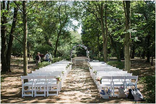 An outdoor ceremony setup photographed from a wide rear angle, showing rows of white folding chairs arranged in two sections along a central aisle in a wooded garden setting. The aisle leads to a floral arch decorated with what appears to be pastel-toned blooms, likely soft pink and lavender flowers. Chair ends are decorated with small floral arrangements in pink and white tones. Two or three people are visible in the background near the arch, appearing to make final setup preparations. The overall styling is soft and romantic with a garden-party aesthetic, using white furniture and pastel florals against the natural tree canopy.