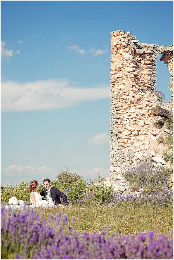 A couple sits together in a field beside the ruins of a tall stone wall, likely in Provence given the blooming purple lavender visible in the foreground. The bride wears a white strapless gown and has auburn hair styled up, while the groom is dressed in a dark suit with a light-colored tie or cravat; they appear to be facing each other in conversation. The wide-angle portrait composition places the couple small within the frame, emphasizing the crumbling limestone ruin tower and open landscape around them. The overall styling is romantic and relaxed, with the natural lavender field and ancient stonework creating a distinctly rustic, southern French aesthetic.