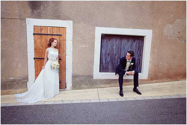 A couple portrait taken outdoors against a rendered stone wall featuring two wooden-doored openings. The bride stands in the left doorway wearing a white one-shoulder gown with a flowing train, holding a small bouquet of white and cream flowers, with auburn hair styled up. The groom sits casually on the ledge of the right recessed window opening, wearing a dark navy suit with a light sage-green tie, looking across at the bride. The two are separated by a gap in the wall, creating a symmetrical, playful composition. The shot is a wide portrait taken at street level, emphasizing the architectural framing and the distance-yet-connection between the two subjects.