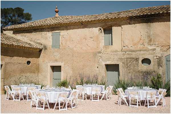 An outdoor wedding reception setup in a gravel courtyard in front of a rustic Provençal farmhouse with aged ochre render, sage green shuttered doors, and terracotta roof tiles. Five round tables are dressed in white linen with glassware already set, surrounded by white folding chairs, arranged in a loose row against a border of lavender and wildflowers. No guests are present, suggesting the photo was taken before the reception began. Wide shot capturing the full scene with the venue building as the dominant backdrop. Potential venue feature image.