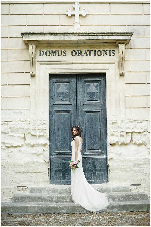 A bridal portrait taken outdoors in front of a historic stone chapel or oratory, with the inscription 'Domus Orationis' and a stone cross above the entrance. The bride stands on the stone steps before a large, weathered dark grey double door, turned slightly to face the camera over her shoulder. She wears a fitted ivory sleeveless gown with a flowing train and holds a bouquet featuring blush pink, coral, and peach blooms with green foliage. Her dark hair is worn down. The composition is a full-length portrait shot, capturing the architectural facade as a strong backdrop against the pale stone building.