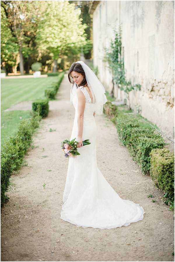 A bridal portrait taken outdoors along a gravel pathway flanked by low trimmed box hedges, set against a stone wall with climbing ivy. The bride stands in a three-quarter rear-facing pose, glancing downward, wearing a fitted ivory lace gown with a low open back, a short two-tiered veil, and holding a compact bouquet of blush pink, mauve, and purple blooms with dark green foliage. The dress has a modest train resting on the gravel path. The overall styling is classic with a romantic, understated feel. Full-length portrait shot with soft natural light.