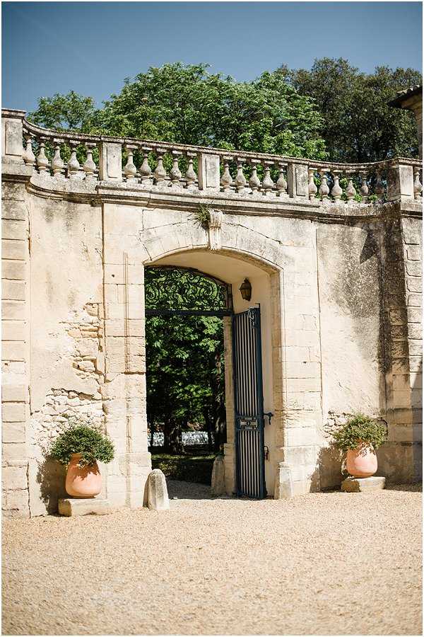 Wide shot of a classical French chateau or domaine entrance gate, featuring a tall arched stone doorway with ornate wrought-iron gate panels painted in dark blue-grey, topped with a decorative ironwork arch. The surrounding limestone wall is crowned with a carved stone balustrade running along the top. Two terracotta urns with clipped round shrubs flank the entrance on a gravel courtyard. A small lantern is mounted on the arch above the gate. No people are visible in the image. Potential venue feature image.