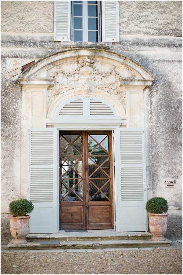 A close-up architectural shot of the entrance facade of a French chateau or manor house, featuring a grand double wooden door with cross-pattern glazing, framed by pale blue-grey louvred shutters and an ornate carved stone pediment with decorative relief detailing above the arch. Two clipped boxwood topiaries in terracotta pots flank the doorway on a stone step. A small sign reading 'Accueil' with a directional arrow is visible to the right of the entrance. The image contains no people and serves as a venue detail shot. Potential venue feature image.
